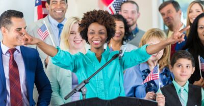 A female African American political candidate addresses supporters at her political campaign launch. She is standing at a podium. Diverse family and friends are gathered around her.