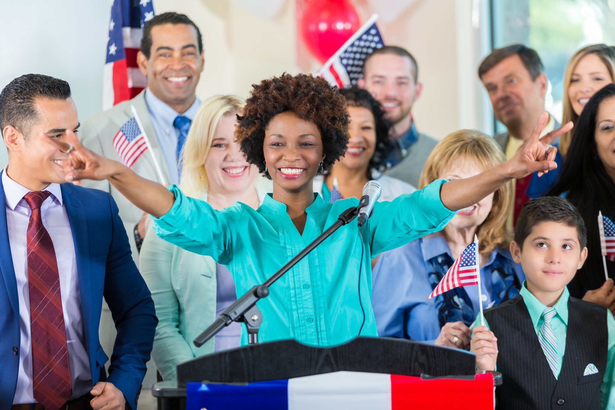 A female African American political candidate addresses supporters at her political campaign launch. She is standing at a podium. Diverse family and friends are gathered around her.