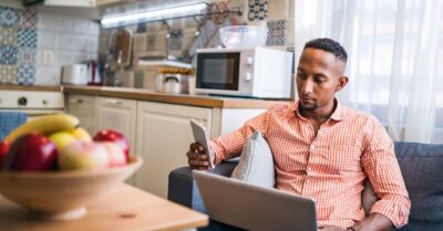 A man holding a cell phone and a laptop participates in phone canvassing from his kitchen at home.