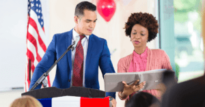 A candidate and staffer are at a podium reviewing notes in a binder.