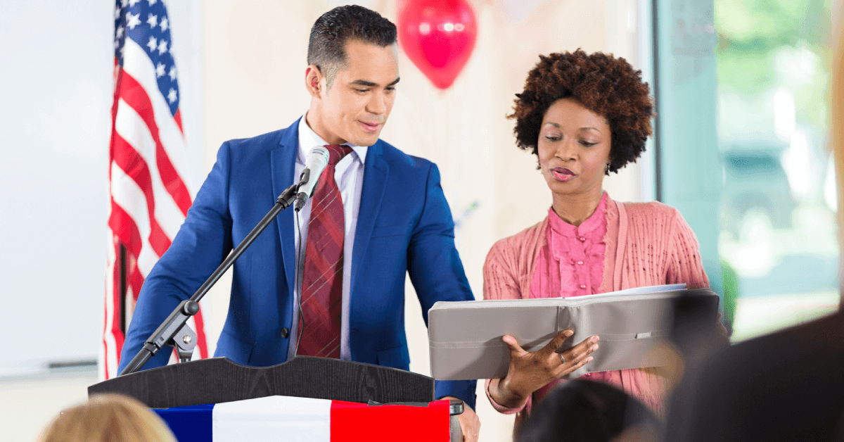 A candidate and staffer are at a podium reviewing notes in a binder.