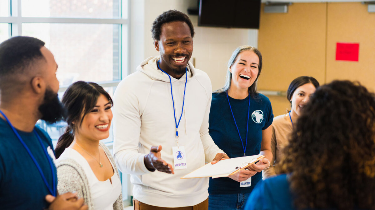 A facilitator leads a discussion in a community meeting, holding a clipboard while speaking to attendees.