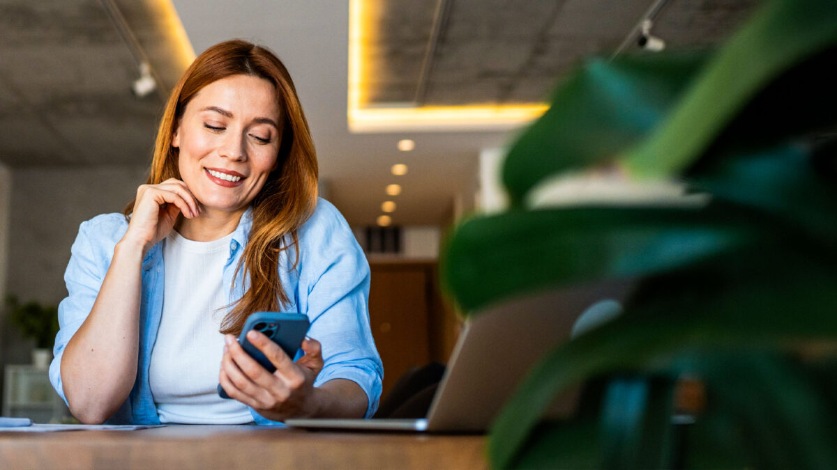 A woman sits at a workspace smiling while checking her phone next to her laptop.