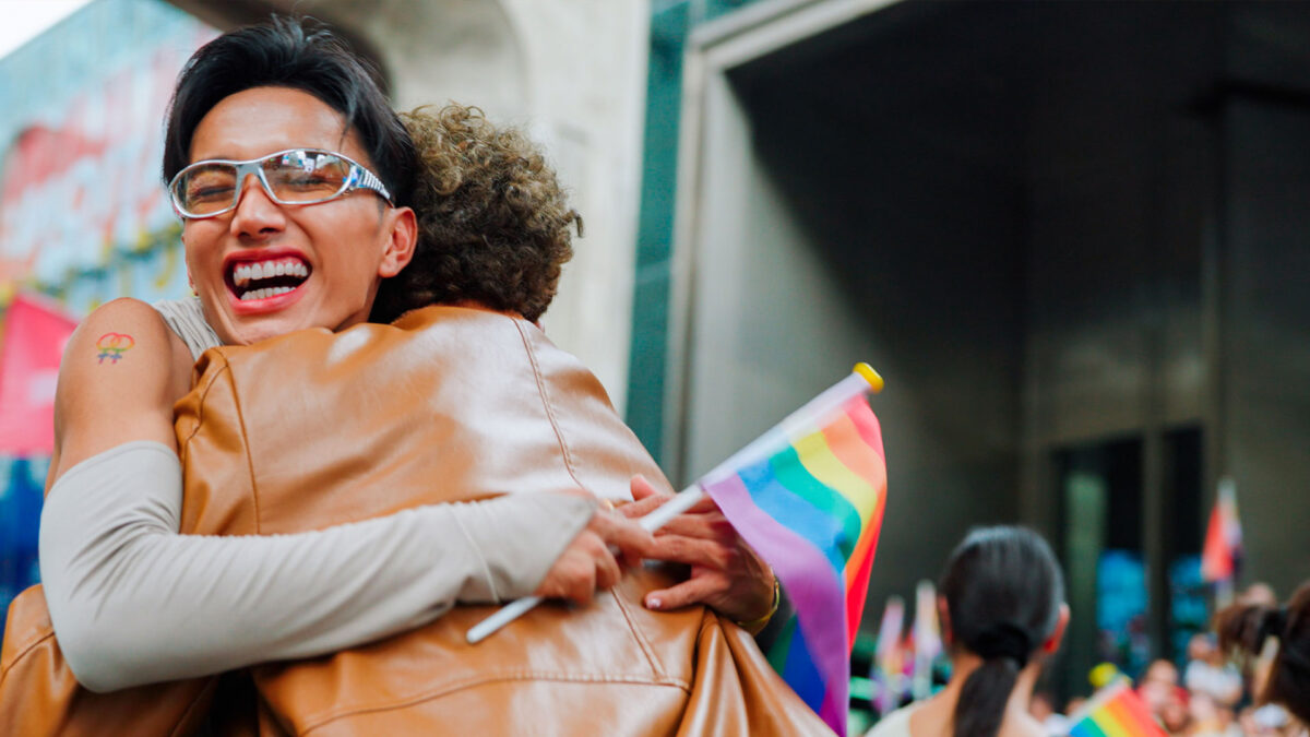 Two people embrace joyfully at a Pride event, holding rainbow flag.