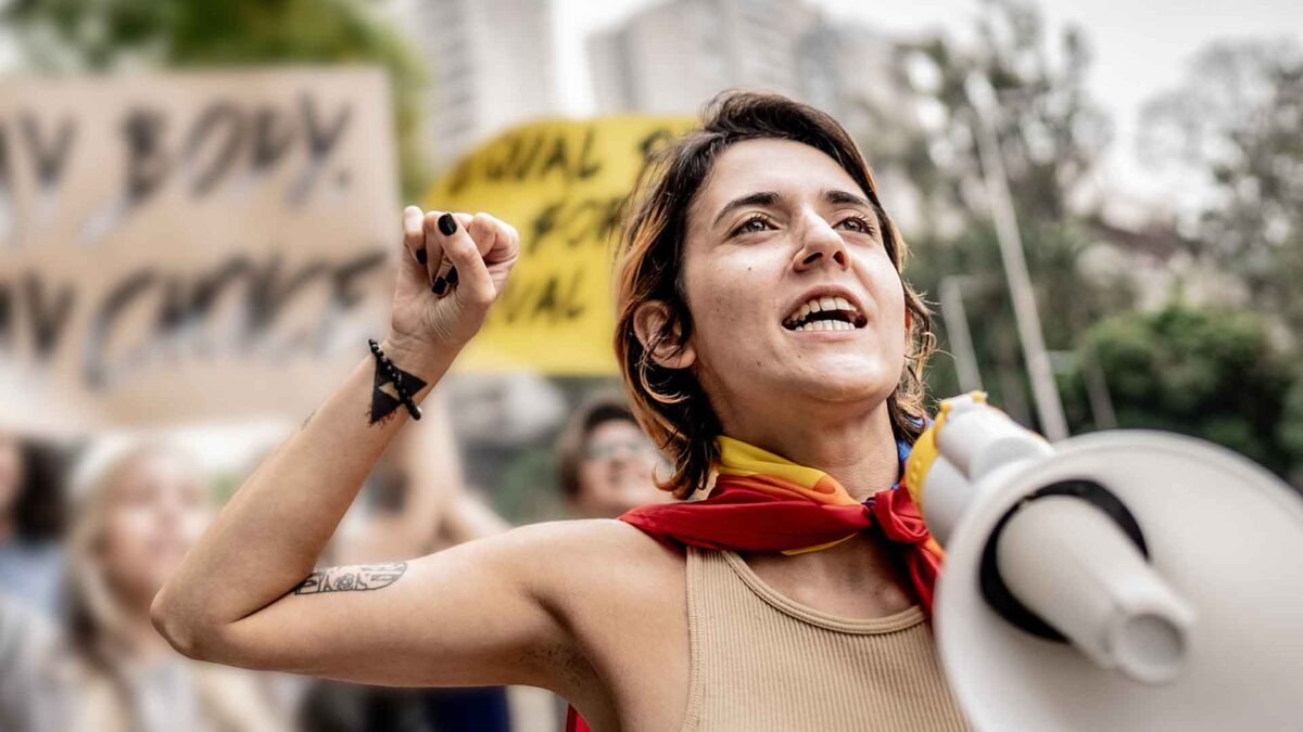 Person speaking into a megaphone at a rally, raising a fist in front of protest signs