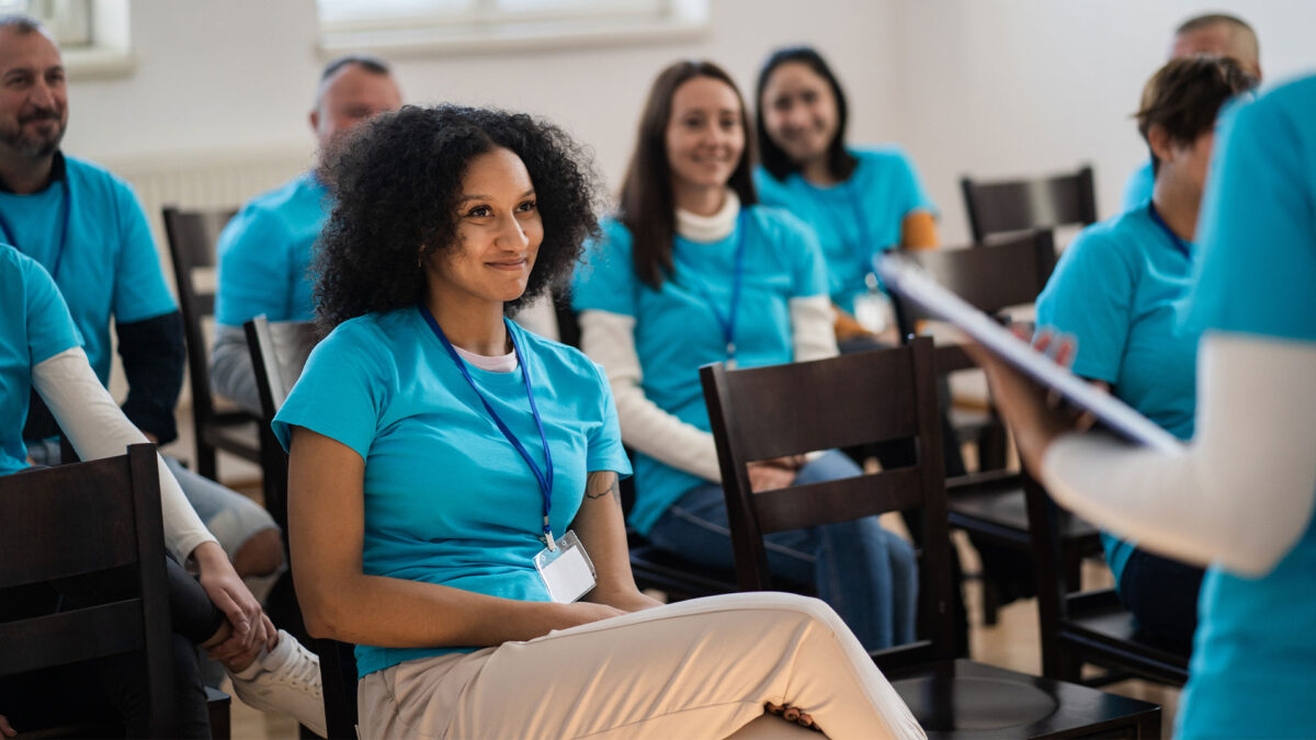 A group of volunteers in matching blue shirts sit in chairs, listening and smiling during a training session.