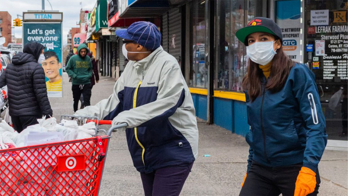 Rep. Alexandria Ocasio-Cortez and volunteers push a red shopping cart full of supplies down a city sidewalk, delivering goods during a community relief effort.