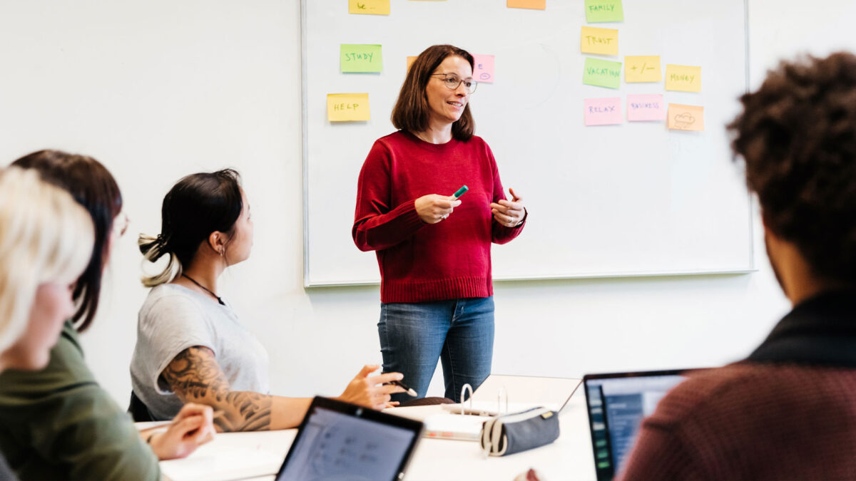 A facilitator stands at the front of a room leading a training for staff and volunteers, with colorful sticky notes covering a whiteboard behind her.