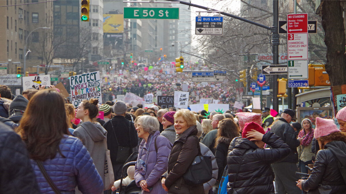 A massive crowd of demonstrators fills a city street during a daytime march, many carrying colorful protest signs.