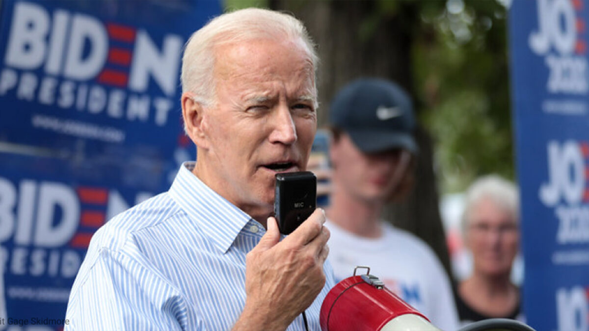 Joe Biden speaks into a microphone at an outdoor campaign rally, with blue campaign signs visible in the background.