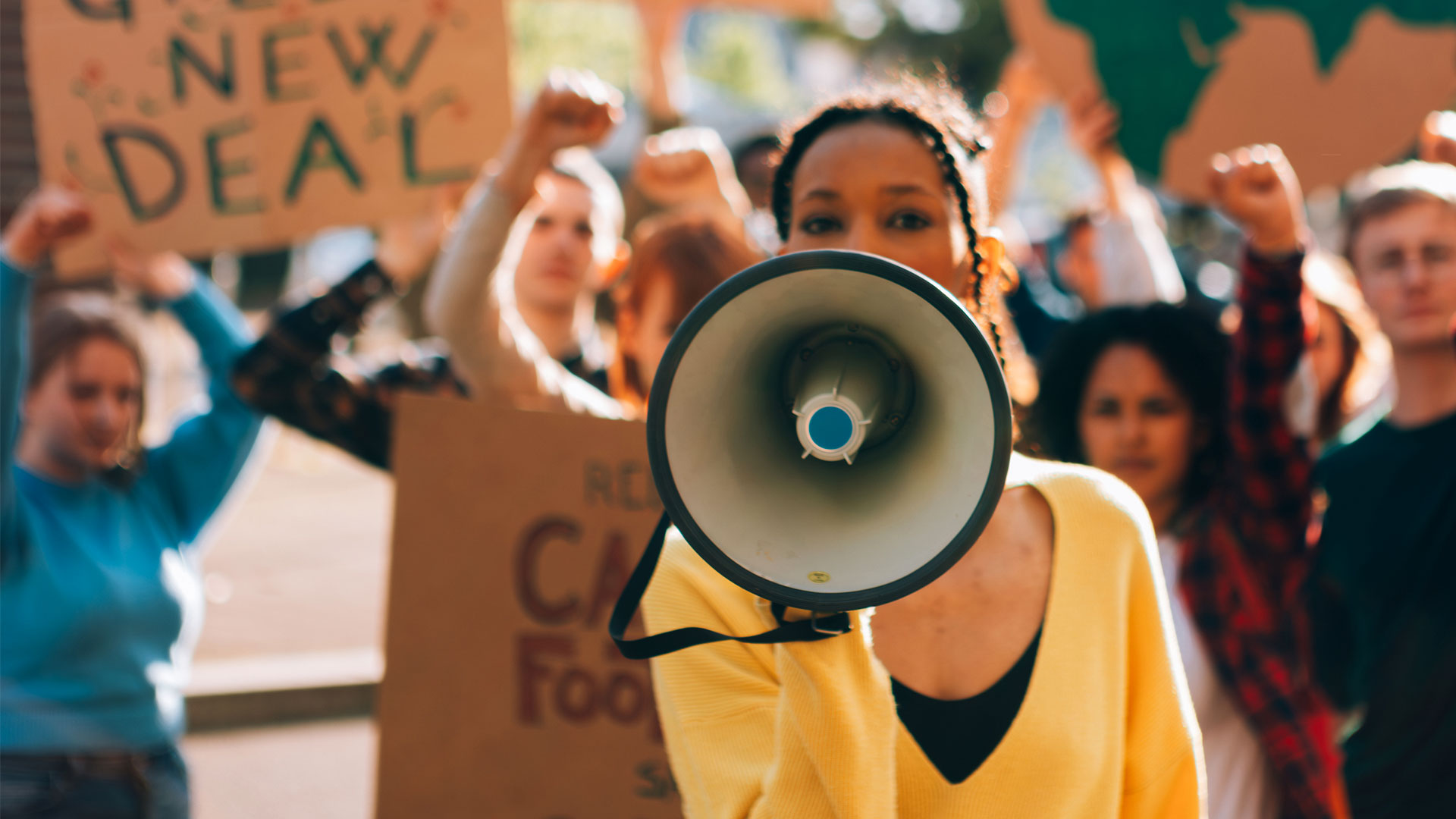 A protester speaking through a megaphone at a rally, surrounded by people holding signs, representing grassroots organizing and collective action.