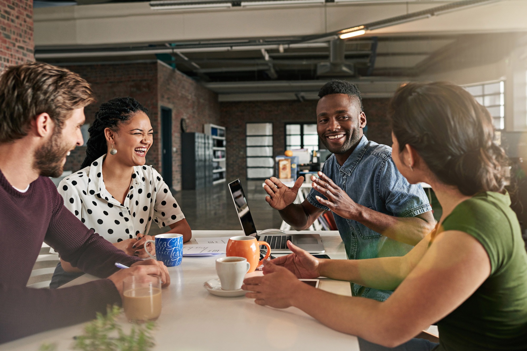 Small team meeting around a table, talking and laughing together in a collaborative workspace.